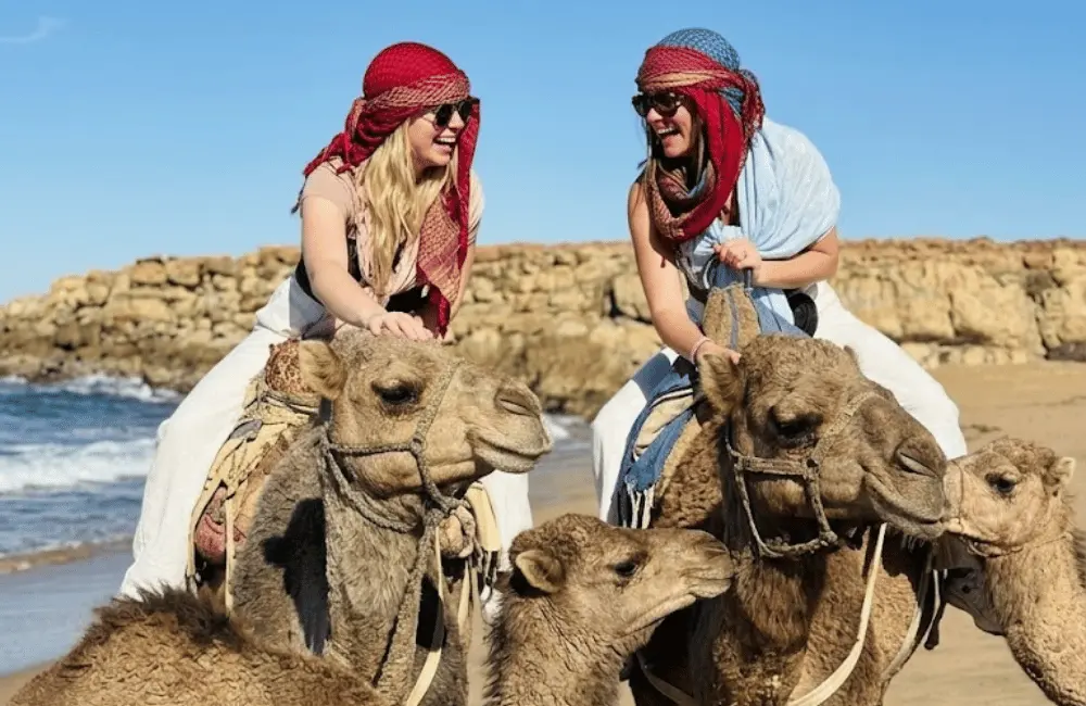 Une rencontre pleine de sourires Deux voyageuses sourient avec plusieurs chameaux près d’une plage rocheuse